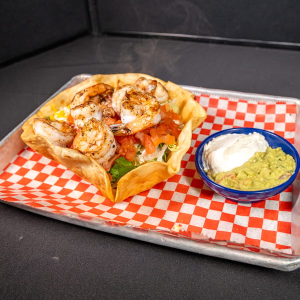 Steaming shrimp taco salad in a fried tortilla bowl with guacamole and sour cream.