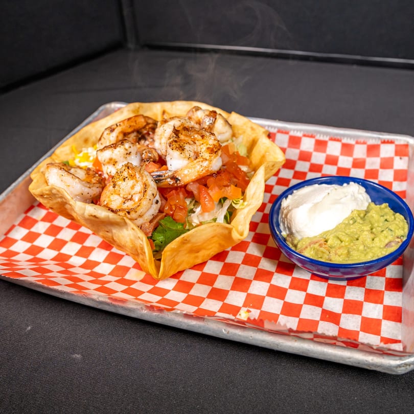 Steaming shrimp taco salad in a fried tortilla bowl with guacamole and sour cream.